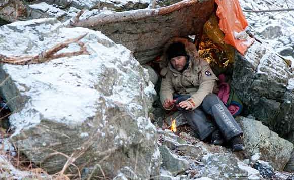 Survivorman Les Stroud under a makeshift shelter with a fire
