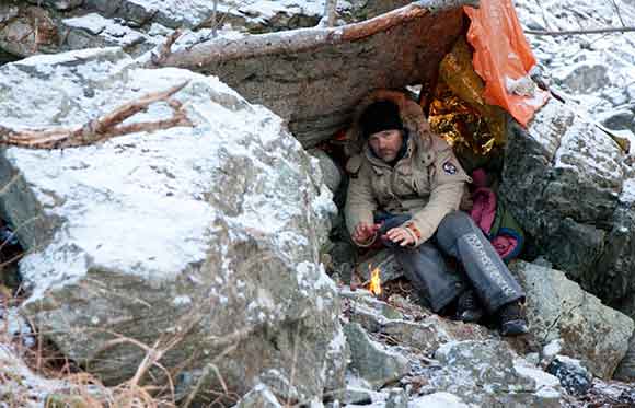 Survivorman Les Stroud under a makeshift shelter with a fire
